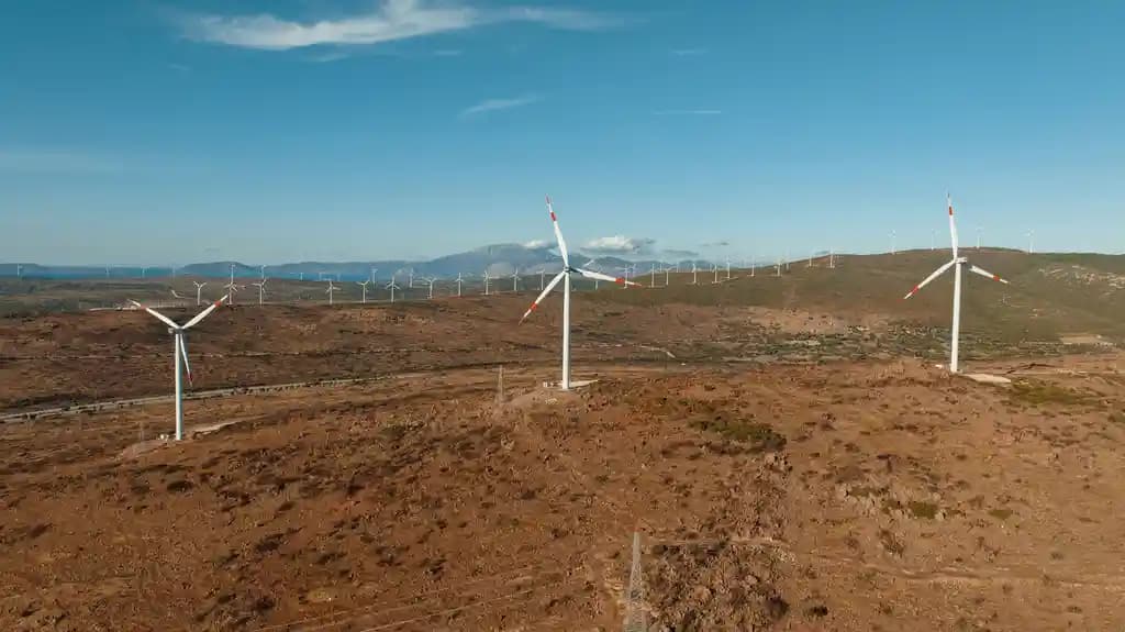 Drone industri terbang mendekati turbin angin raksasa di ladang pembangkit listrik.